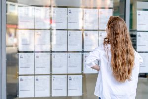 A woman with long hair stands in front of a glass window, reviewing job listings that are displayed on the wall outside a public space.