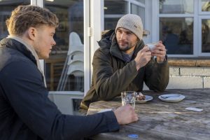 Medium shot of two male friends sitting down, having a coffee, outside a cafe at Druridge Bay in Northumberland. They have been walking the dogs along the beach while wearing casual, warm clothing, as it is in winter. They are looking at each other with serious expressions as they are talking about their mental well-being.
