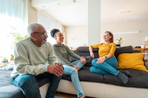 A mixed race family is gathered in a bright living room, smiling and engaged in a casual chat. The group includes a male and female adult, and a young woman, all dressed in comfortable casual wear.